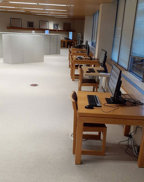 Square wooden tables line a wall with windows. A matching chair sits with each desk. On top of each desk is a computer monitor, keyboard, and mouse.