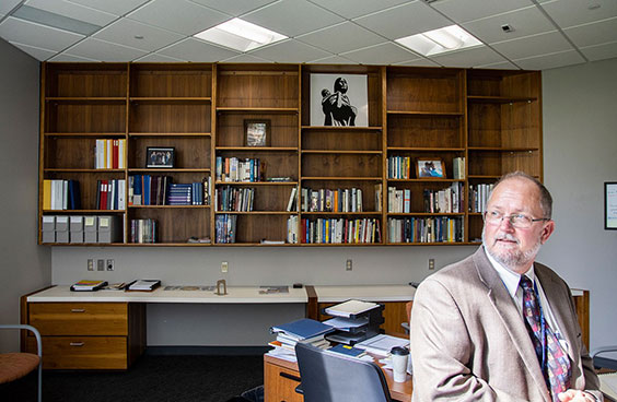 A man wearing glasses and a gray suit stands looking to the side with a very large, mostly empty, bookcase behind him