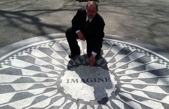 A man in a suit bends down on one knee and uses one hand to reach out and touch a gray and white decorative circle on the ground that says IMAGINE in the middle