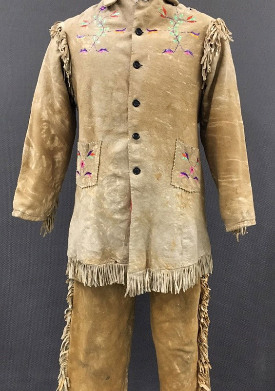 Tan buckskin jacket and pants with fringe on the bottom and shoulders of the jacket and sides of the pants. Colorful floral designs are on the front of the shoulders and pockets of the jacket.
