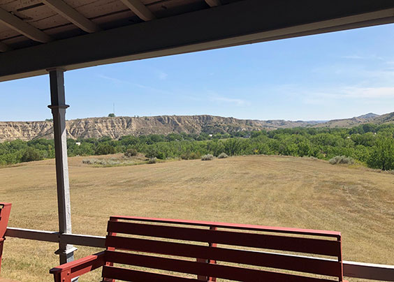 A view looking from a porch with a bench out to a large mowed area with buttes in the background