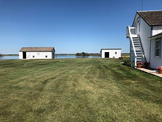 An outdoor view of a large area of mowed lawn with three white buildings and a lake in the background