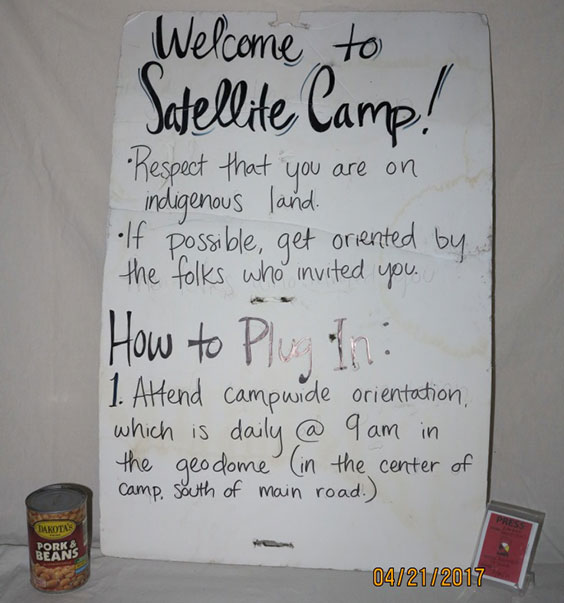 Sign reading Welcome to Satellite Camp! Respect that you are on indigenous land. If possible, get oriented by the folks who infited you. Hot to Plug In: Attend campwide orientation, which is daily @ 9 am in the geodome (in the center of camp, south of main road.)