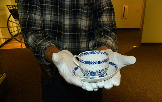 A man in a plaid shirt and white gloves holds a white teacup and saucer with blue flowers and leafy vines