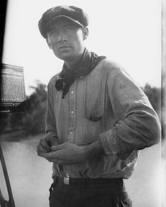 A man stands outdoors wearing a hat, bandana around his neck, button up shirt, and pants with belt.
