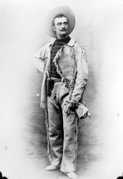 A man with a moustache pointed at the ends stands wearing a cowboy yeat, jacket with tassles, striped shirt, and pants with tassles down the sides.