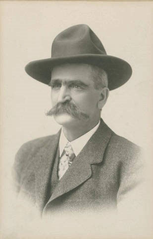 Man with a large mustache wearing a hat and suit complete with vest and tie.