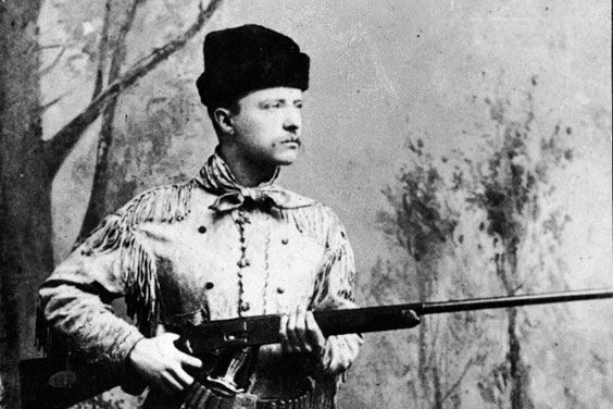 A man holds a gun wearing a beanie looking hat, a jacket with tassles, and a bandana tied around his neck. Trees are in the background.