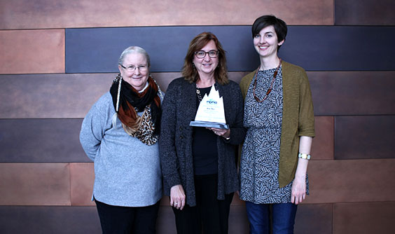 Three women standing in front of copper wall. The middle woman is holding an award.