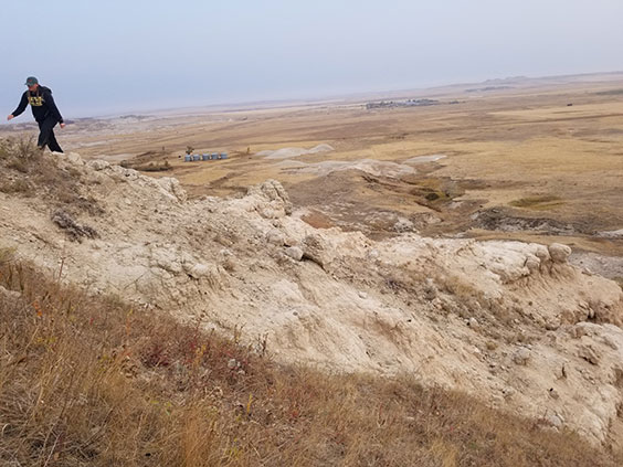 A women wearing a dark colored sweatshirt, pants, and hat hikes up a very brown hill with brown fields shown in the distance behind her