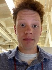 A young man with redish brown hair wearing a white tshirt and collared blue button up shirt