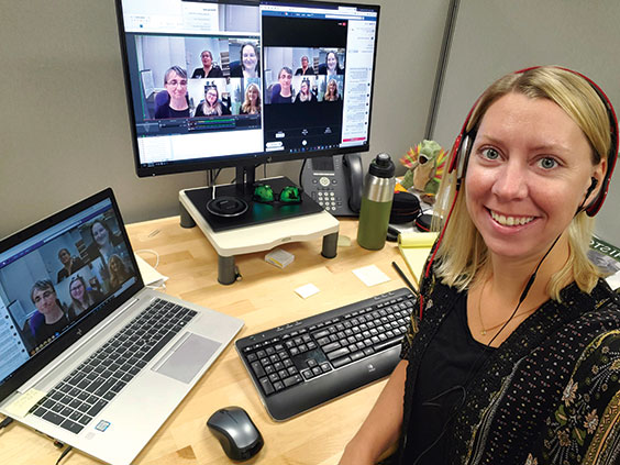 A woman sits at a desk wearing two different headphones with a laptop and another computer monitor running Teams and Facebook Live. Also on the desk are a computer mouse, keyboard, sunglasses, water bottle, telephone, stuffed t rex, and other odds and ends.