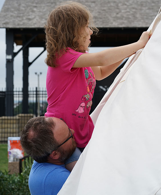 An adult man holds up a young girl while she helps assemble a tipi