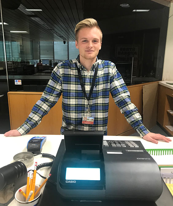 College-aged man standing by a cash register ready to greet people