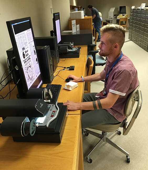 College-aged man sitting at a computer looking at microfilmed newspaper from the microfilm machine next to him